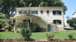 A two-story house with a stone staircase leading to a balcony, surrounded by a well-maintained lawn and a swimming pool.