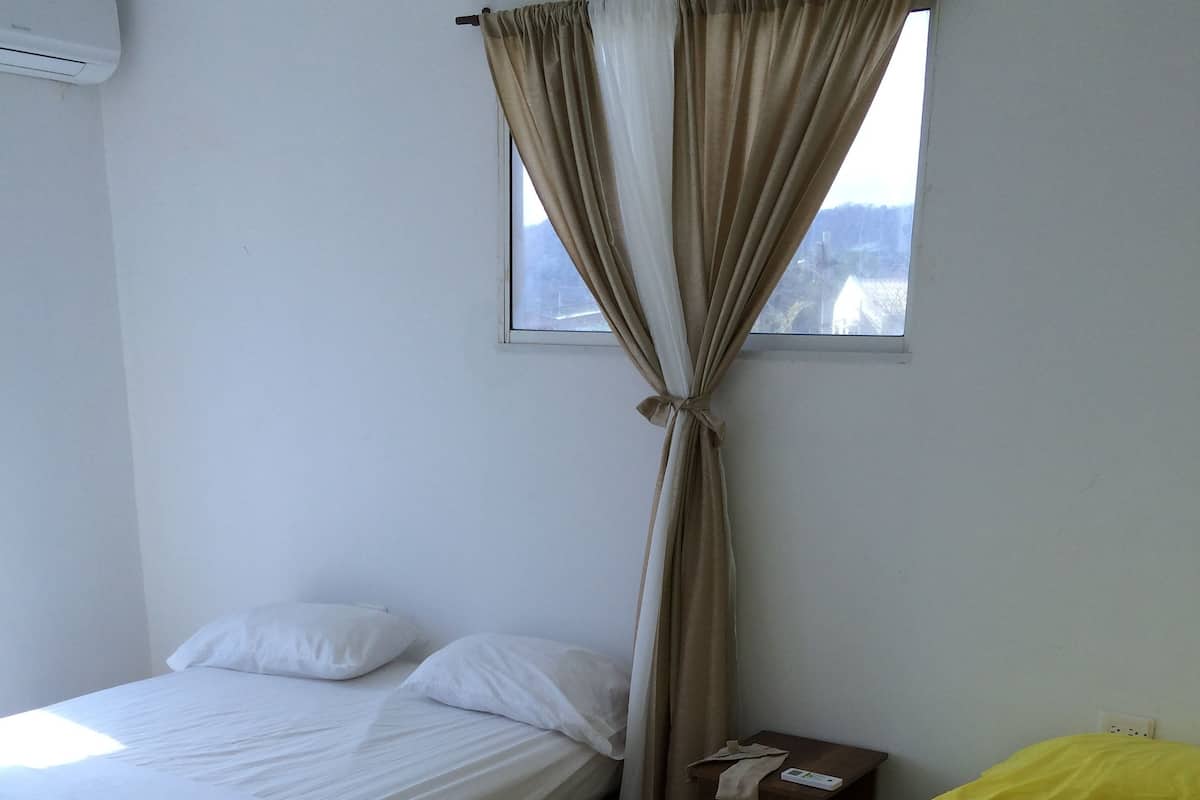 A room with two beds, one with white bedding and the other with yellow bedding, a window with curtains, and a bedside table with a lamp.