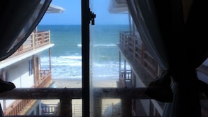 Vista desde una ventana con cortinas, que da a una playa con estructuras de madera y una barandilla.