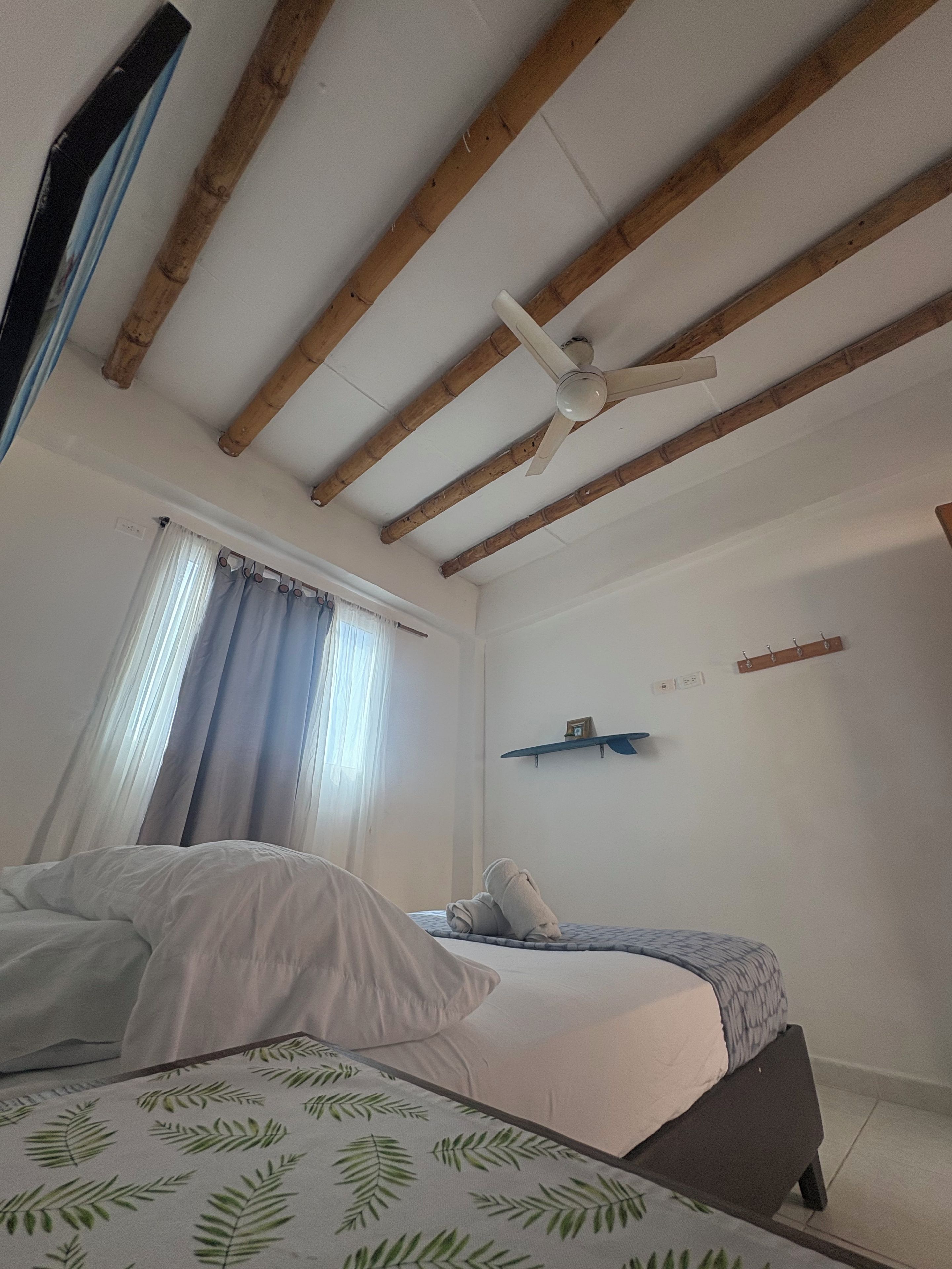 Un dormitorio con dos camas, vigas de madera en el techo, un ventilador de techo, una ventana con cortinas y un estante montado en la pared.