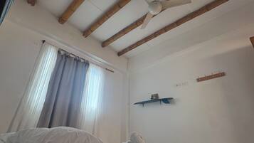 Un dormitorio con dos camas, vigas de madera en el techo, un ventilador de techo, una ventana con cortinas y un estante montado en la pared.