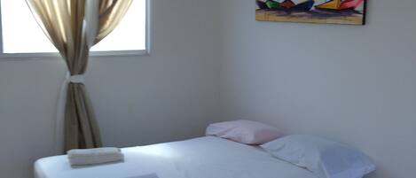 A room with a bed, a small bedside table, a window with curtains, an air conditioning unit, and a painting on the wall.