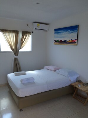 Una habitación con cama, una mesita de noche, ventana con cortinas, aire acondicionado y un cuadro en la pared.