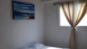 Una cama individual con ropa de cama blanca, una mesita de noche amarilla, un cuadro de un paisaje marino y una ventana con cortinas color beige.