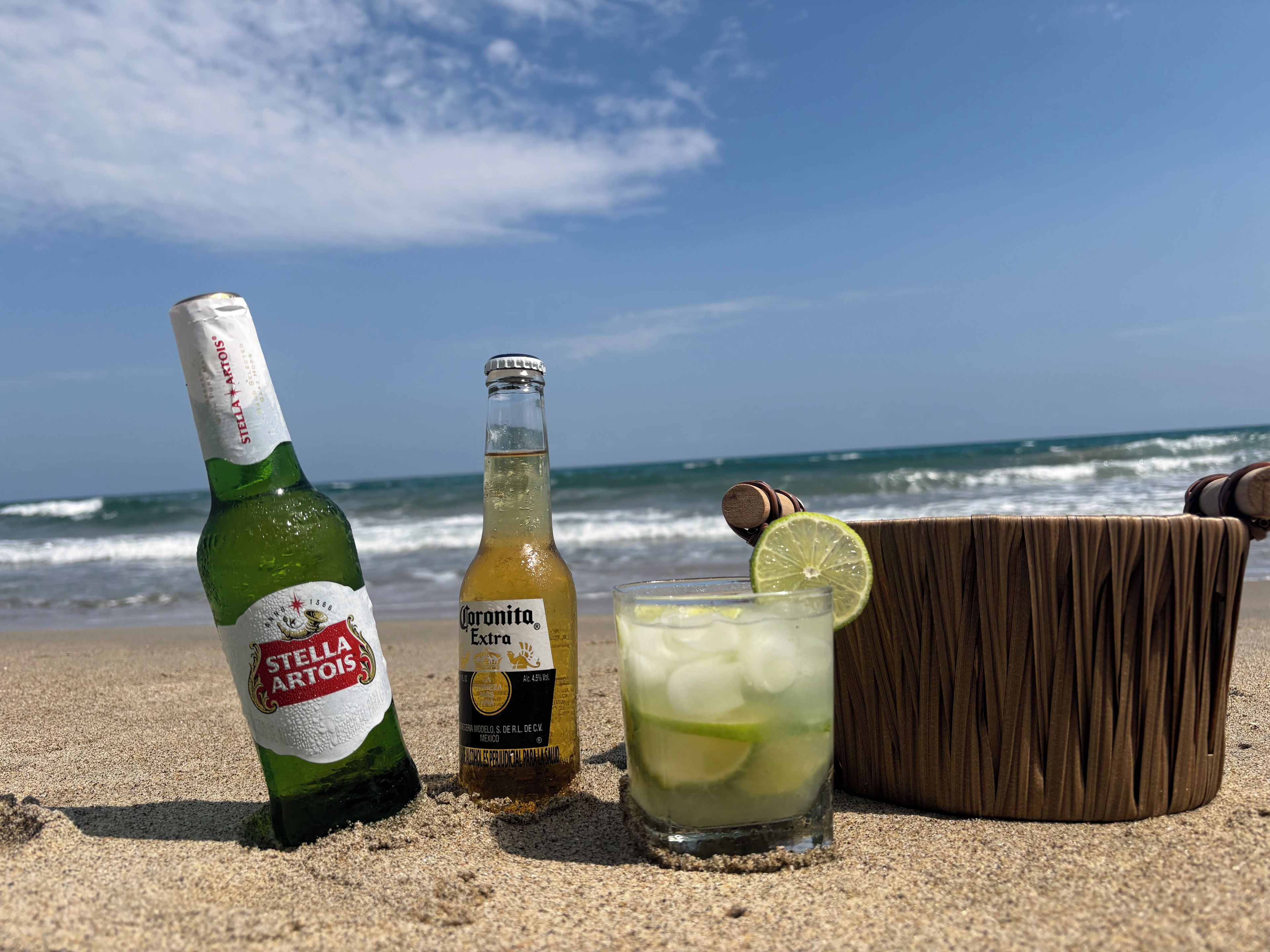 Una botella de cerveza Stella Artois, una botella de cerveza Corona Extra, y una cerveza con una rodaja de lima en una playa arenosa con el océano al fondo.