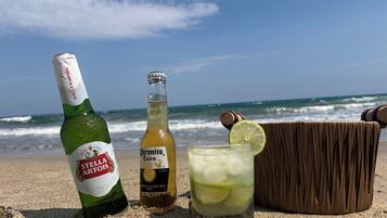 Una botella de cerveza Stella Artois, una botella de cerveza Corona Extra, y una cerveza con una rodaja de lima en una playa arenosa con el océano al fondo.