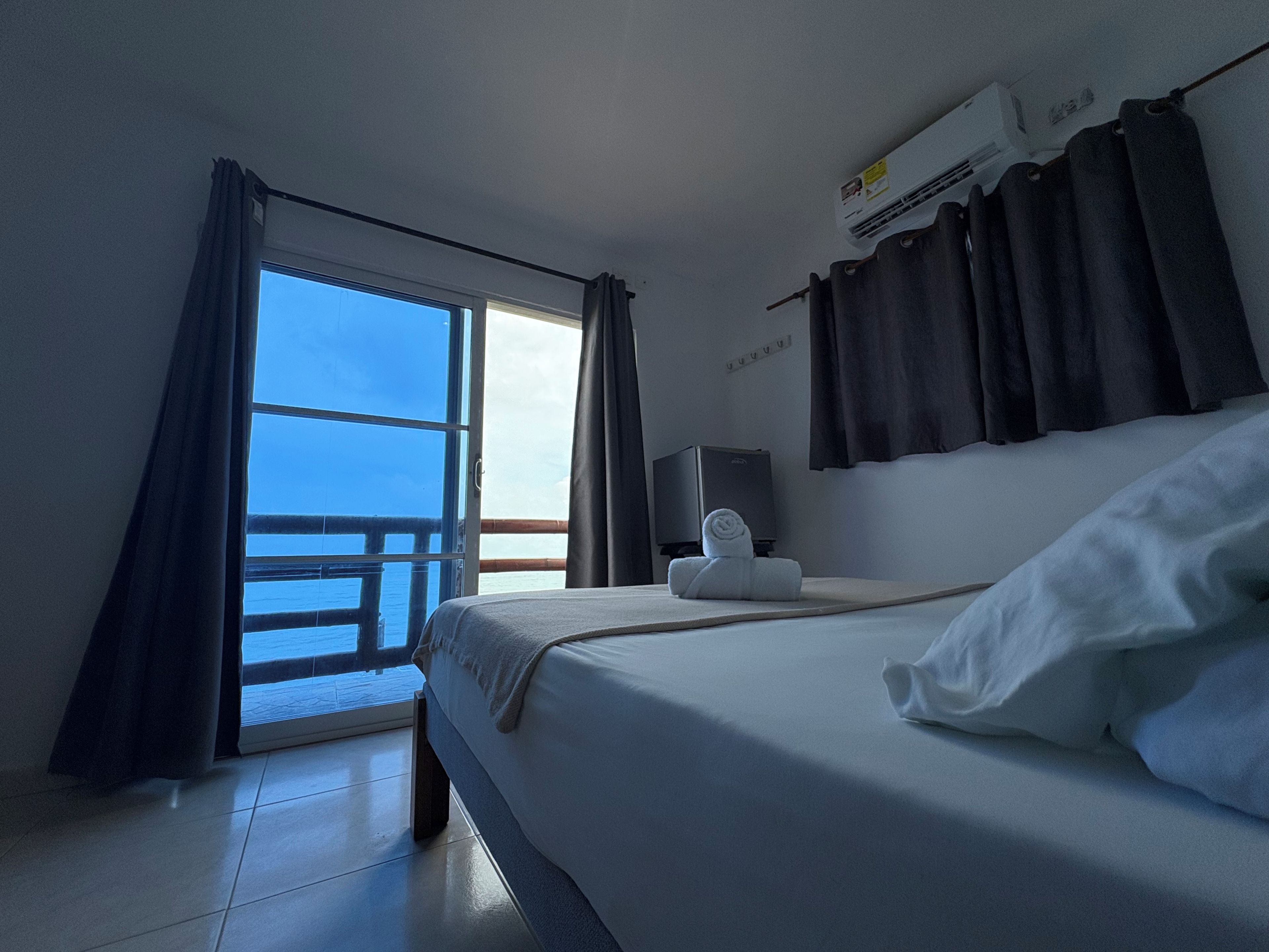 A hotel room with a bed, a TV, a towel, and a view of the sea through a large window.