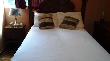 Una cama bien hecha con dos almohadones estampados, una mesita de noche con lámpara y un buró con teléfono.