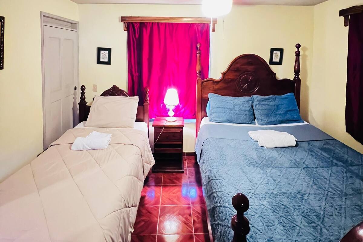 A room with two beds, a ceiling fan, a red curtain, and a nightstand with a lamp.