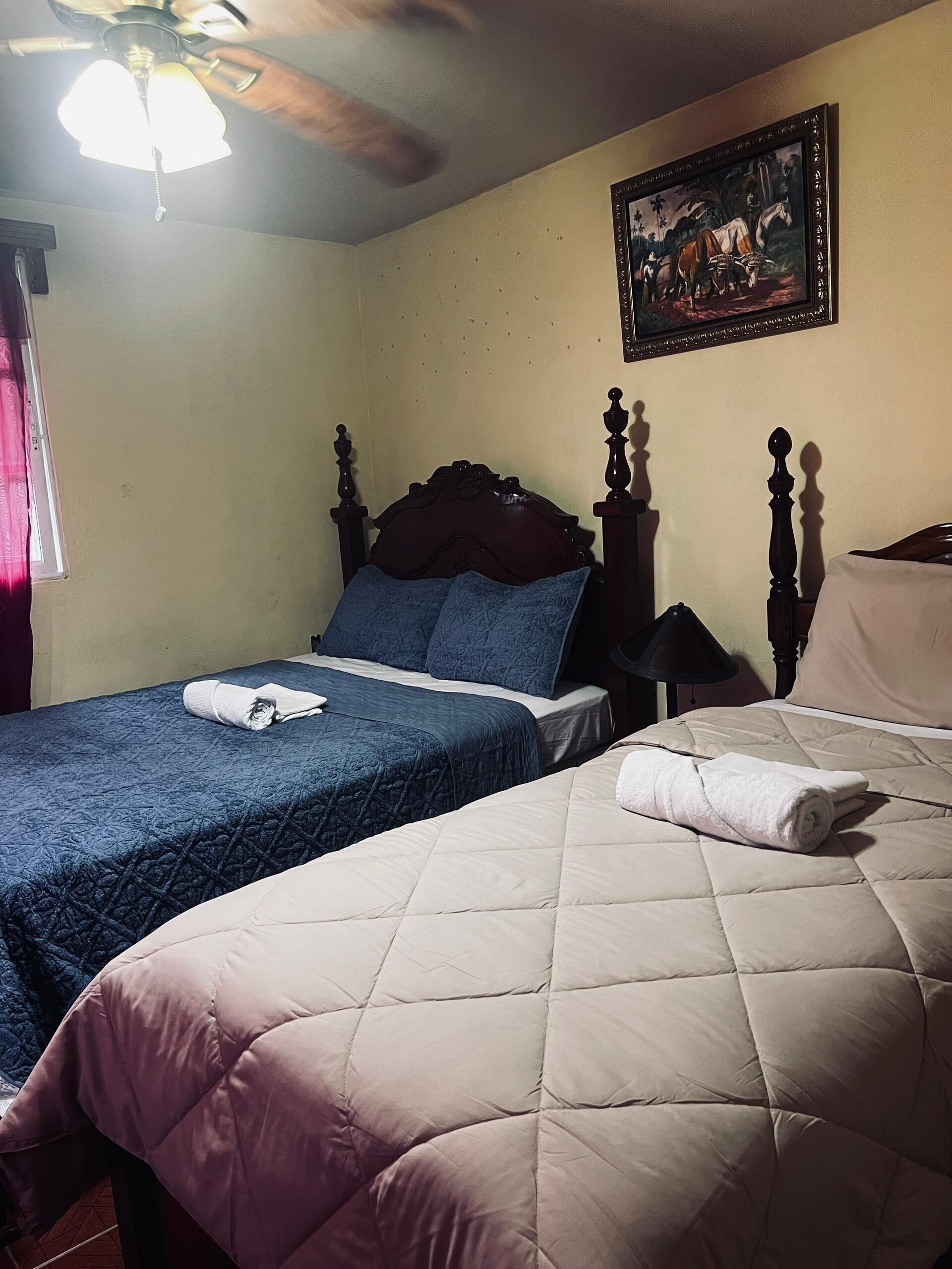 Habitación de hotel con dos camas, una con colcha azul y la otra con colcha color beige. Hay un cuadro en la pared y un ventilador de techo sobre las camas.