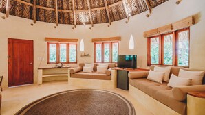 A spacious living area with a thatched roof, a sofa set, a television, and large windows offering a view of greenery. - One Santuario Natural (Dibulla)