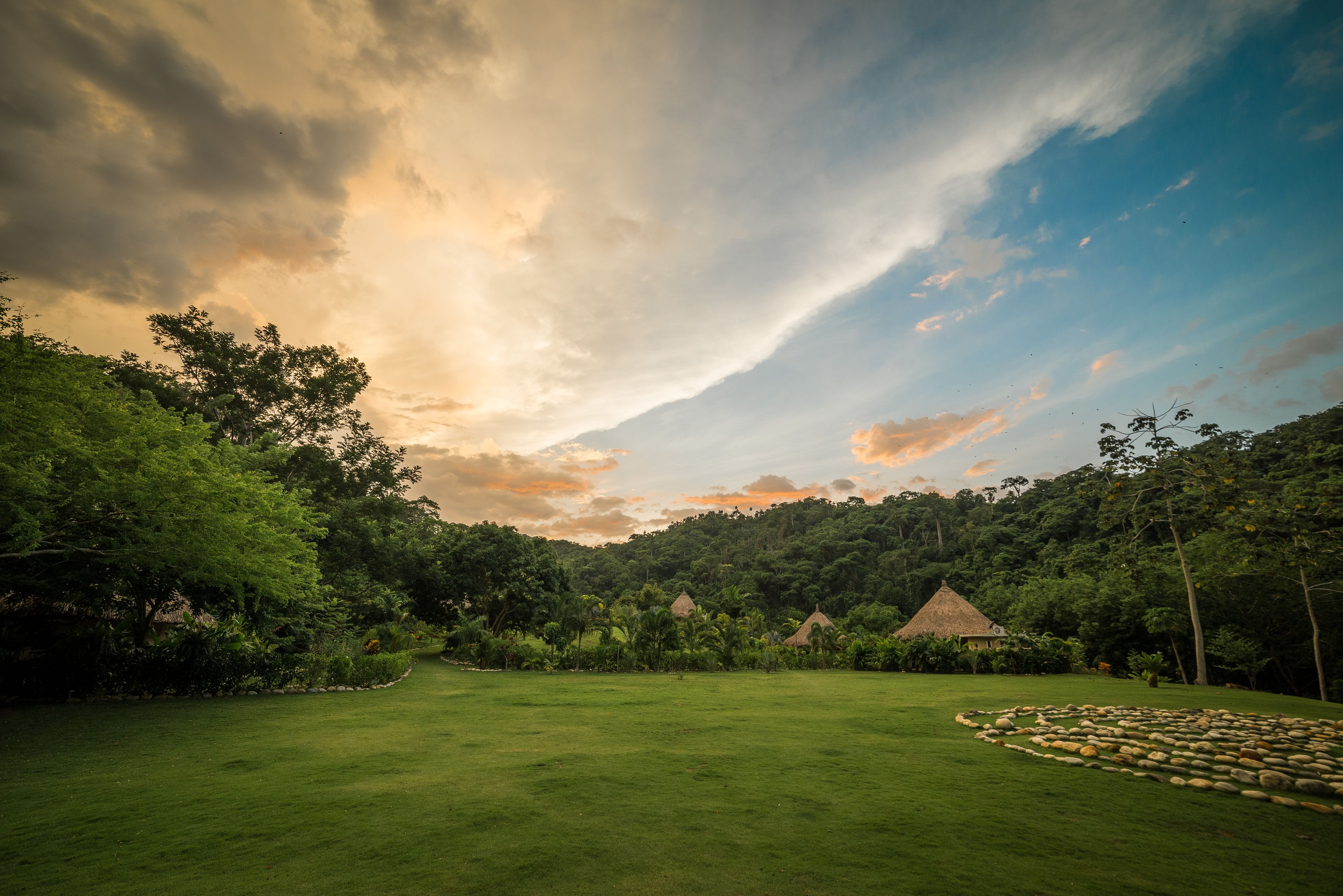 A grassy field with a thatched-roof hut and another hut in the background, surrounded by dense forest.