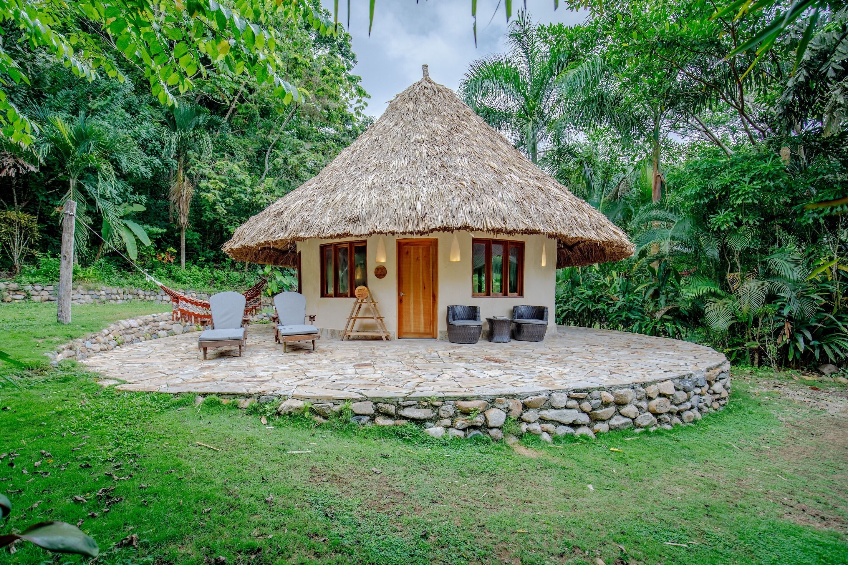 Una cabaña con techo de paja y patio empedrado de piedra, rodeada de exuberante follaje.