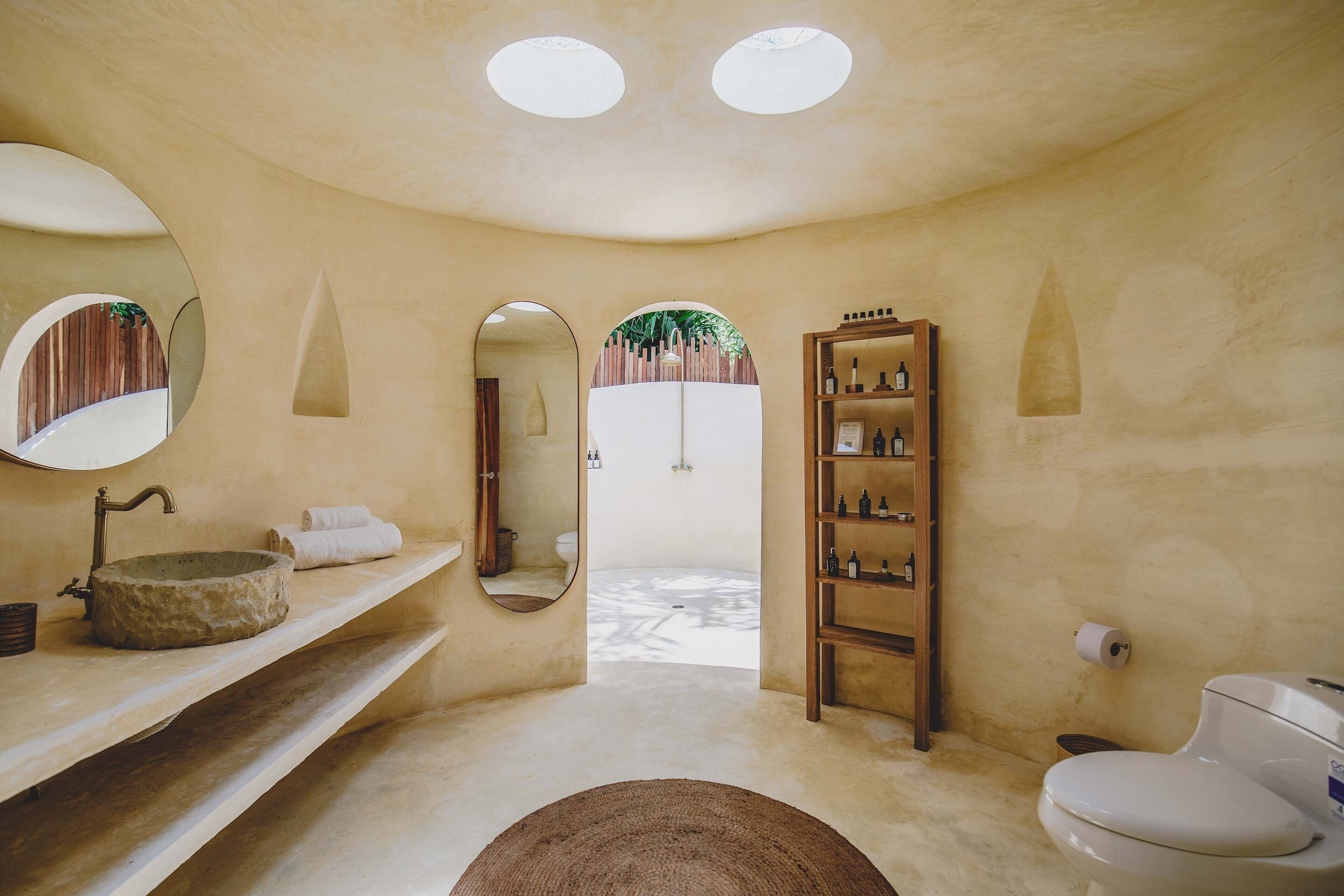 A bathroom with a stone sink, a wooden shelf with toiletries, a toilet, and a mirror.
