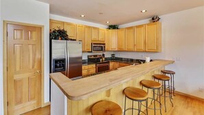 A modern kitchen with wooden cabinets, a stainless steel refrigerator, a microwave, an oven, and a wooden bar with stools. - Pines 101 Ski in out 4 BR Town House (Breckenridge)