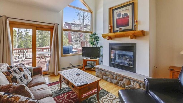 A living room with a fireplace, television, and a large window overlooking a deck. - Pines 101 Ski in out 4 BR Town House (Breckenridge)