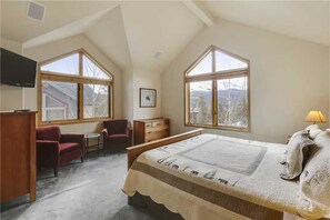 Ein Schlafzimmer mit einem großen Bett, zwei Fenstern, einem Fernseher, einem Schreibtisch und einem Stuhl.