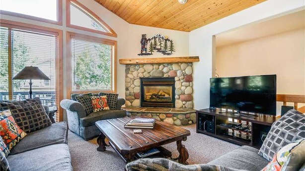 A cozy living room with a stone fireplace, a flat-screen TV, and a large window with blinds. - Windward Way Retreat House (Steamboat Springs)