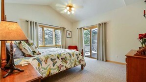 A bedroom with a bed, a desk, a lamp, a ceiling fan, and large windows with curtains. - Windward Way Retreat House (Steamboat Springs)