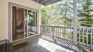 A covered outdoor area with a wooden deck, a sliding glass door leading to a room, and a view of trees. - Windward Way Retreat House (Steamboat Springs)