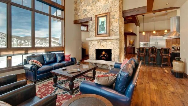 A spacious living room with a stone fireplace, leather sofas, and large windows offering a view of the mountains. - Rocky Fells House (Breckenridge)