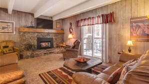 A cozy living room with a stone fireplace, comfortable seating, and a large window with curtains. - Columbine 105 Condo (Breckenridge)