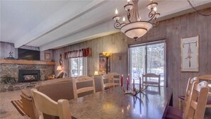 A dining area with a large table, wooden chairs, a chandelier, a fireplace, and a large window. - Columbine 105 Condo (Breckenridge)