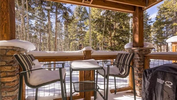 A covered outdoor seating area with stone walls and metal chairs, overlooking a snowy forest. - Columbine 105 Condo (Breckenridge)