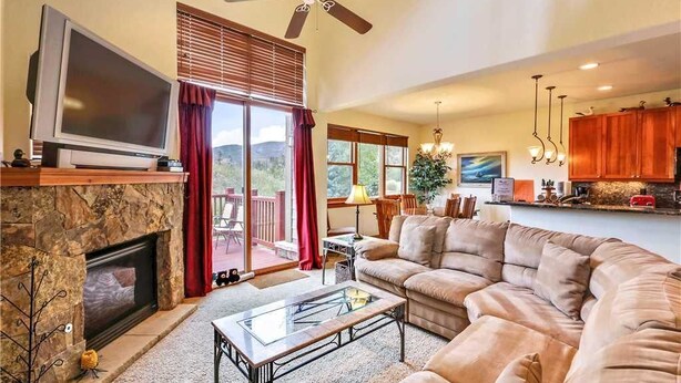 A living room with a fireplace, a large sectional sofa, a television, and a dining area with a view of the outdoors. - Highland Green 91 Town House (Breckenridge)