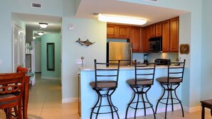 A kitchen with a central island, three bar stools, wooden cabinets, a stainless steel refrigerator, and a microwave. - 302 Seychelles (Panama City Beach)