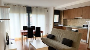 Un espacio habitable compacto con sofá, una mesa de comedor con sillas, una zona de cocina con armarios y electrodomésticos, y un ventanal con cortinas.