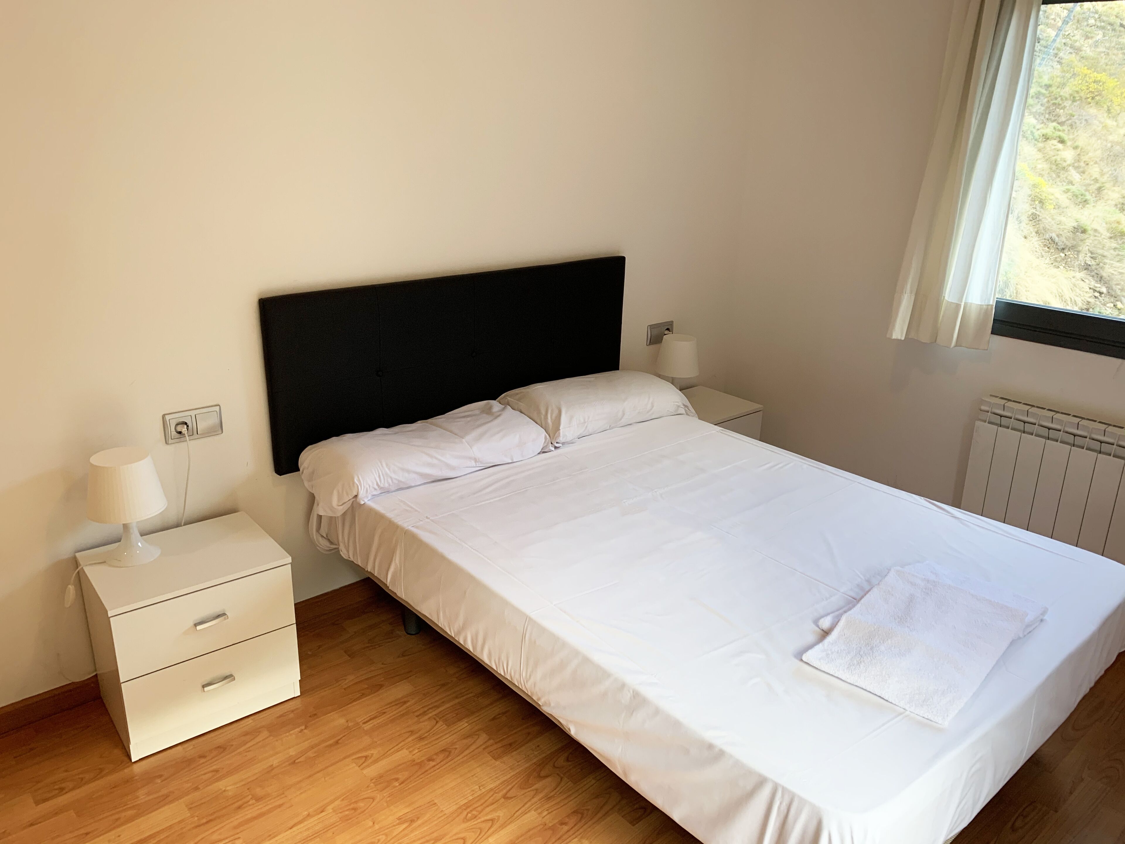 A neatly made bed with white linens, a bedside table with a lamp, and a window with curtains.