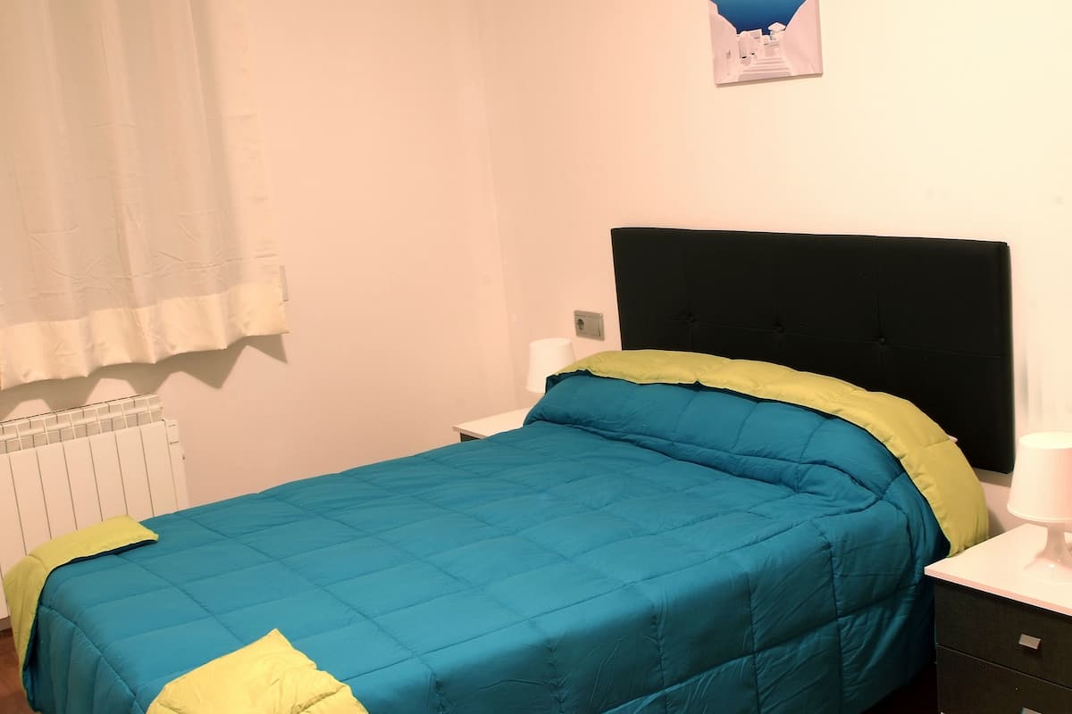 Una cama individual con colcha azul y verde, cabecera negra, mesita de noche blanca y mesita con lámpara.