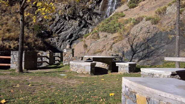 Un área de picnic de piedra con bancos y un muro de piedra, rodeada de césped y árboles.