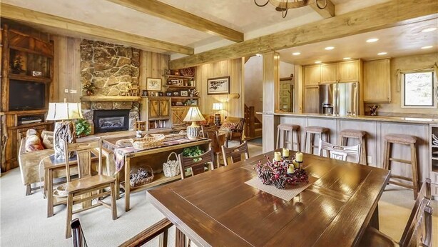 A spacious living room with a large wooden dining table, a stone fireplace, and a kitchen area with wooden cabinets and a bar. - Storm Meadows Townhouse 17 Town House (Steamboat Springs)