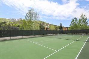 Un court de tennis en plein air, doté d’un filet et entouré d’une clôture, avec en arrière-plan des arbres et des bâtiments.