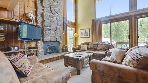 A cozy living room with a stone fireplace, wooden walls, and large windows offering a view of the outdoors.