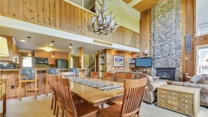 A spacious dining area with a long wooden table and chairs, a stone fireplace, and a cozy living room with a sofa and a television.