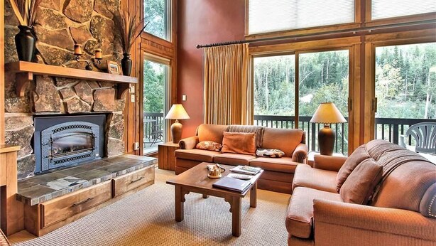 A cozy living room with a stone fireplace, a sofa, a coffee table, and large windows with curtains. - Storm Meadows Townhouse 24 Town House (Steamboat Springs)