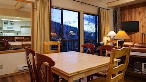 Een eetgedeelte met een houten tafel en stoelen, een televisie aan de muur en uitzicht op het landschap door grote ramen.
