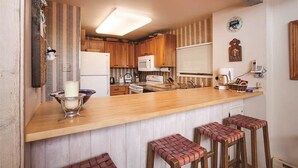 A kitchen with wooden cabinets, a white refrigerator, a microwave, and a wooden bar counter with stools. - Storm Meadows Townhouse 4 Town House (Steamboat Springs)