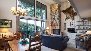 A spacious living room with a stone fireplace, a large window overlooking a forest, and a cozy seating area. - Storm Meadows Townhouse 4 Town House (Steamboat Springs)