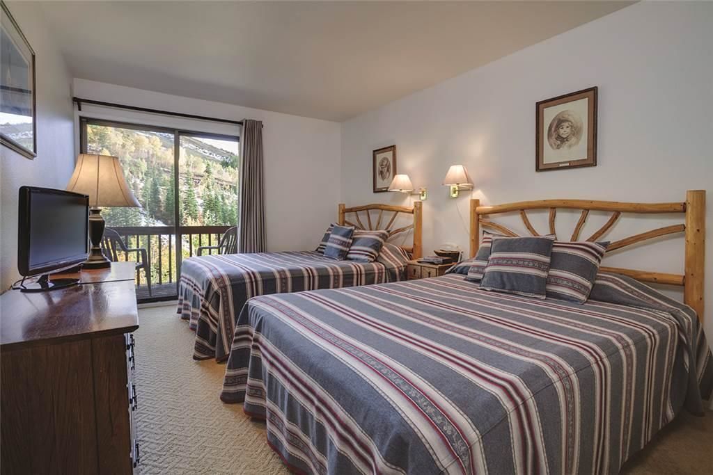 A hotel room with two beds, a TV, a desk, and a balcony with a view of trees.