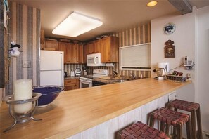 A kitchen with wooden cabinets, a white refrigerator, a microwave, and a wooden countertop. There are bar stools and a blue bowl on the counter.