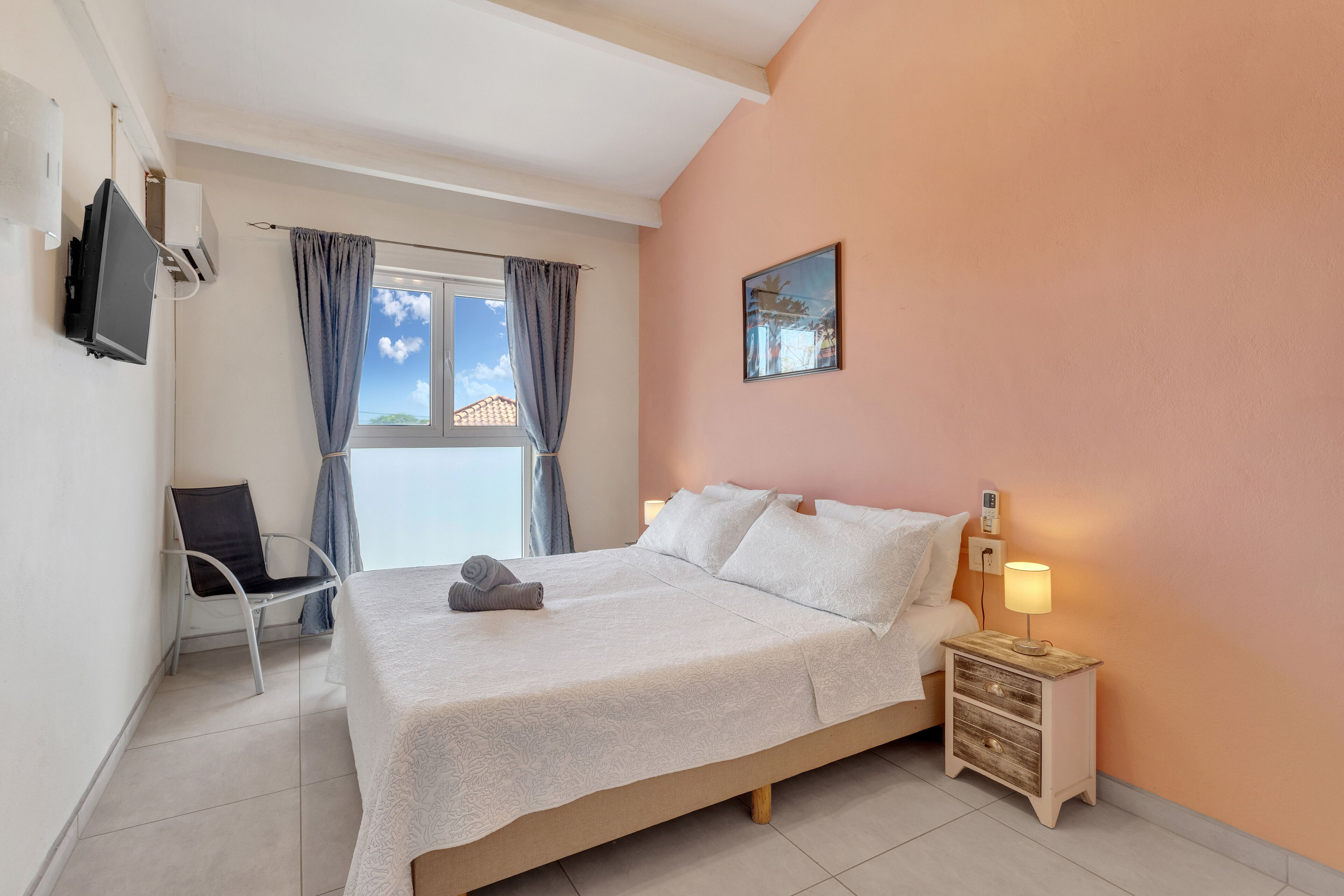 A bedroom with a bed, a chair, a bedside table with a lamp, a TV, and a window with curtains.