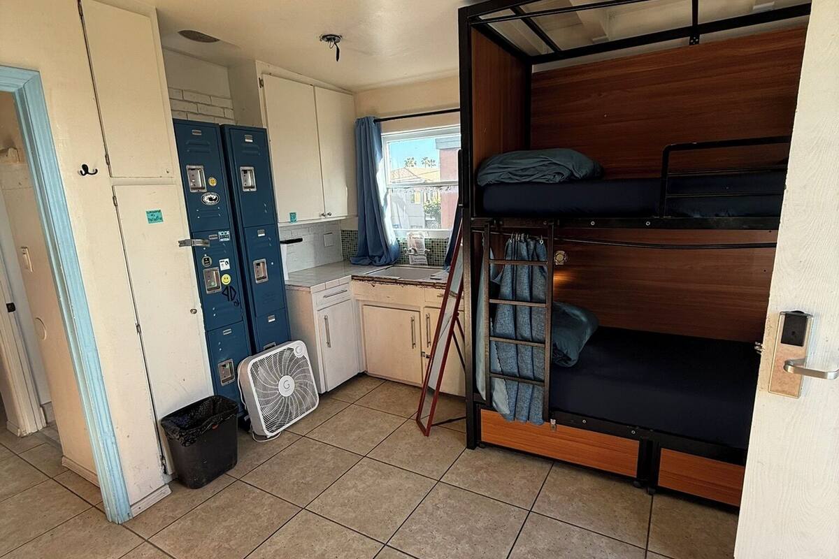 Une petite pièce comprenant un lit superposé, un coin cuisine, un ventilateur et une poubelle.