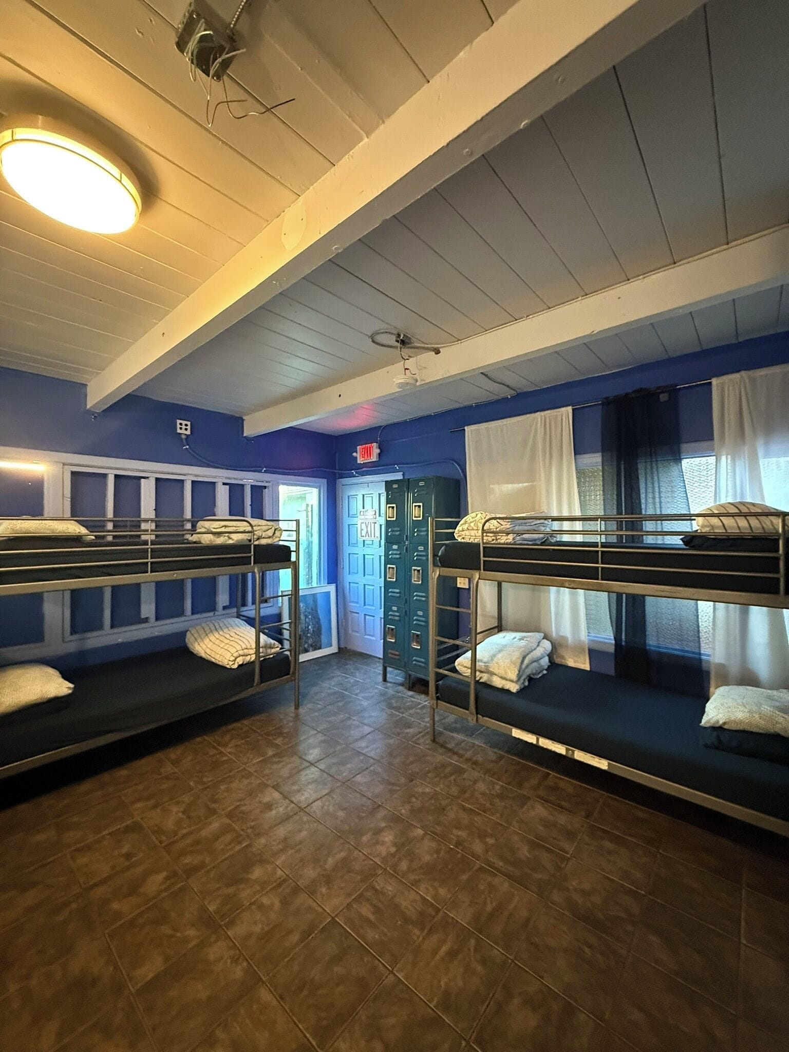 A dormitory room with bunk beds, a door marked 'EXIT,' and a ceiling light fixture.