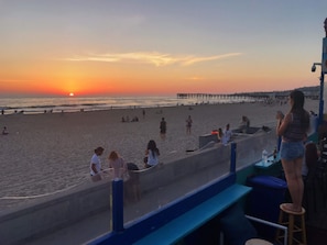 Una playa con un atardecer, gente relajándose y un muelle al fondo.