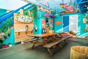 Un espacio exterior animado para comer, con mesa y bancos de madera, rodeado de plantas en macetas y una pared colorida con tablas de surf y adornos.