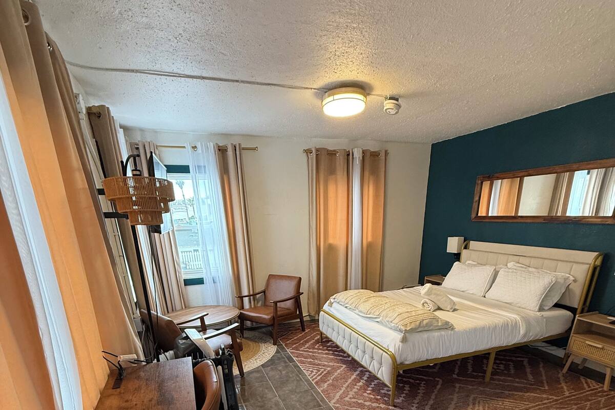 Une chambre d’hôtel avec un lit, un bureau, une chaise, une lampe et une fenêtre avec des rideaux.